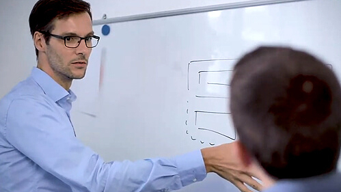 Mann erklärt einem Kollegen den Inhalt an einem Whiteboard