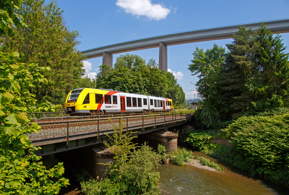 Eine weiß-gelbe HLB Bahn fährt über eine Brücke, die in einem Wald über einen Fluss führt.