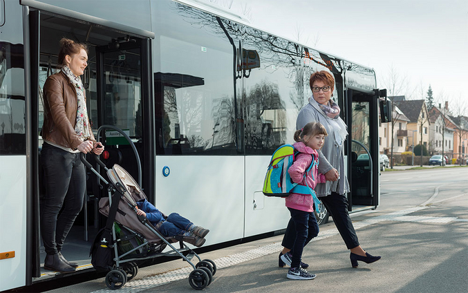 Zwei Frauen, eine davon scheibt einen Buggy mit einem Kleindkind, sowie ein Kind im Vorschulalter verlassen einen modernen Stadtbus