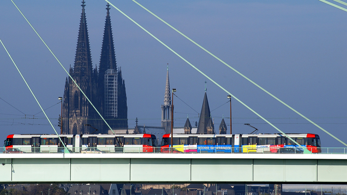 Die Kölner Straßenbahn fährt auf einer Brücke mit dem Kölner Dom im Hintergrund.